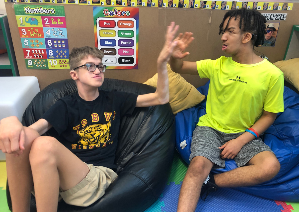 Dos adolescentes chocan los cinco sentados en sillones puff en un aula.