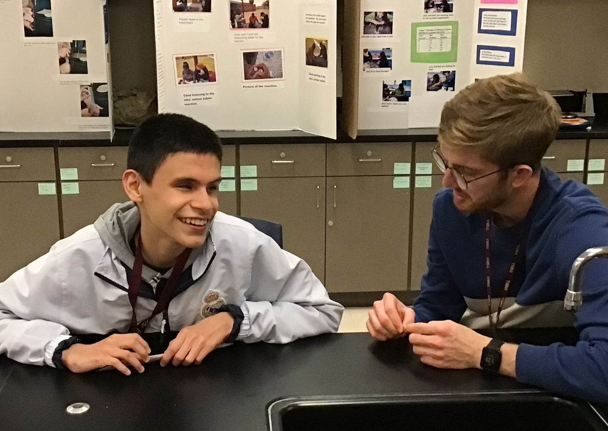 Un adolescente y un adulto sentados en un laboratorio de ciencias con los tableros de la feria de ciencias colocados detrás de ellos.
