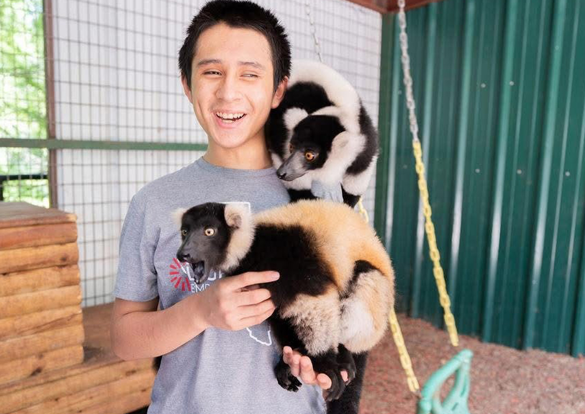 Un estudiante sonríe mientras sostiene un lémur en su hombro y en sus brazos.
