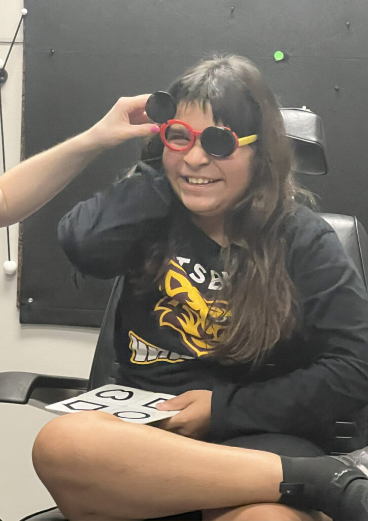 An adolescent wears glasses that cover one eye at a time while an adult moves the occluder. On her lap, the student holds a sheet of testing materials containing drawings of shapes in black outline.