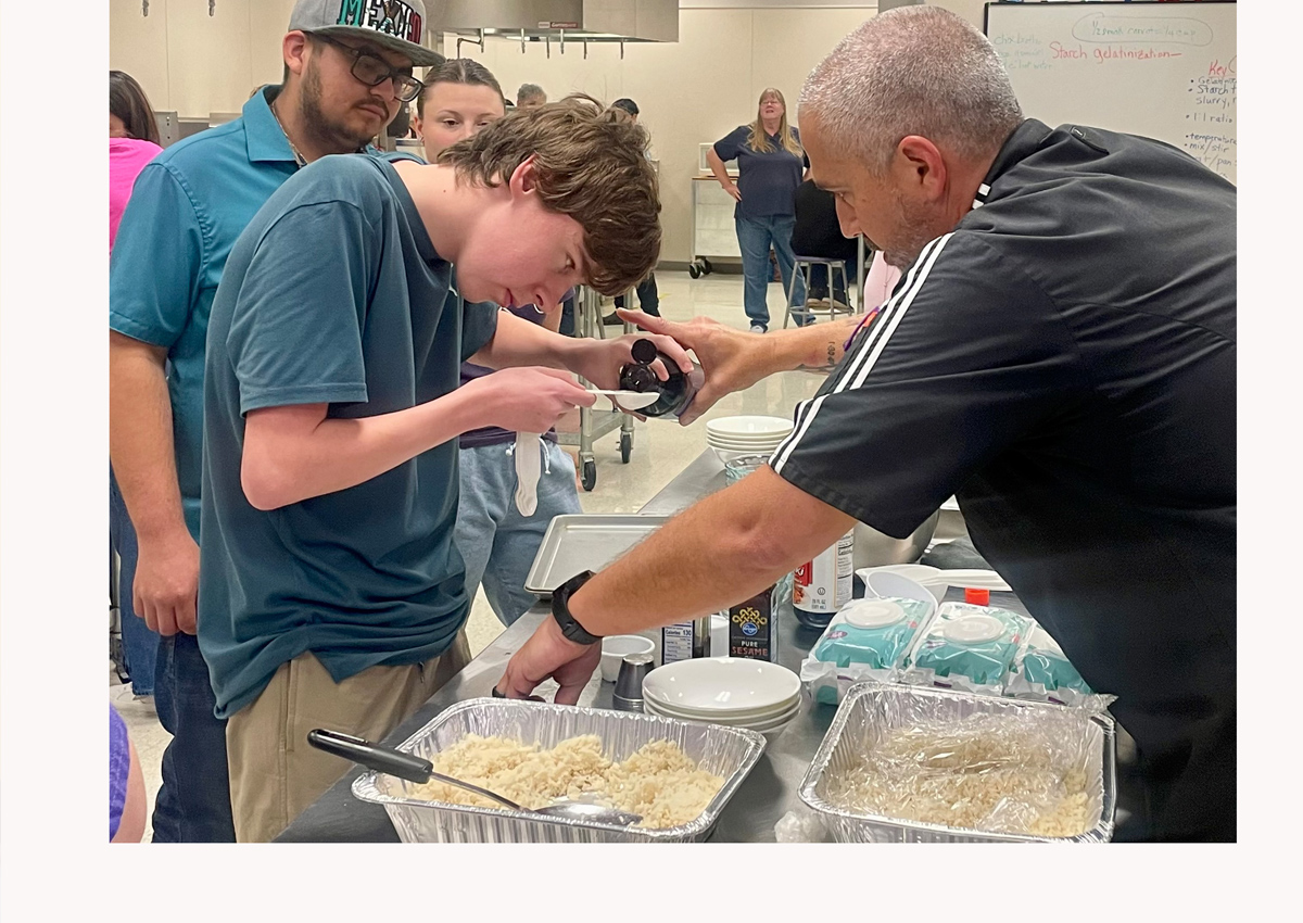 A student pours vanilla into a measuring spoon with support from his instructor.