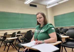 Una joven lee un libro en braille mientras está sentada en un aula con asientos en fila y tres pizarras donde se han dibujado varios pentagramas musicales.