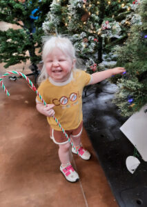 Una niña sonríe mientras toca un árbol de Navidad con la mano izquierda y sujeta un gran bastón de caramelo con la mano derecha