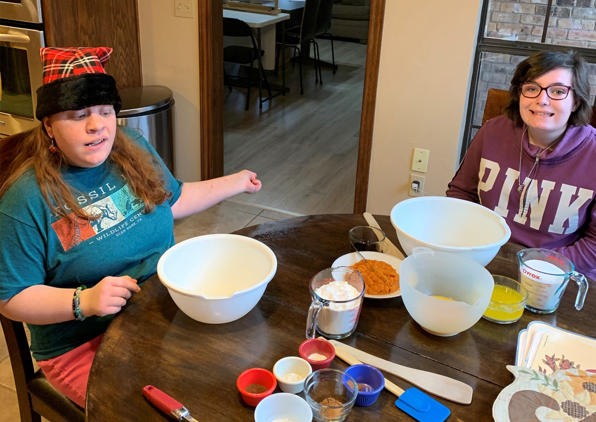 Two young adults sit at a table with mixing bowls and cooking utensils in front of them.