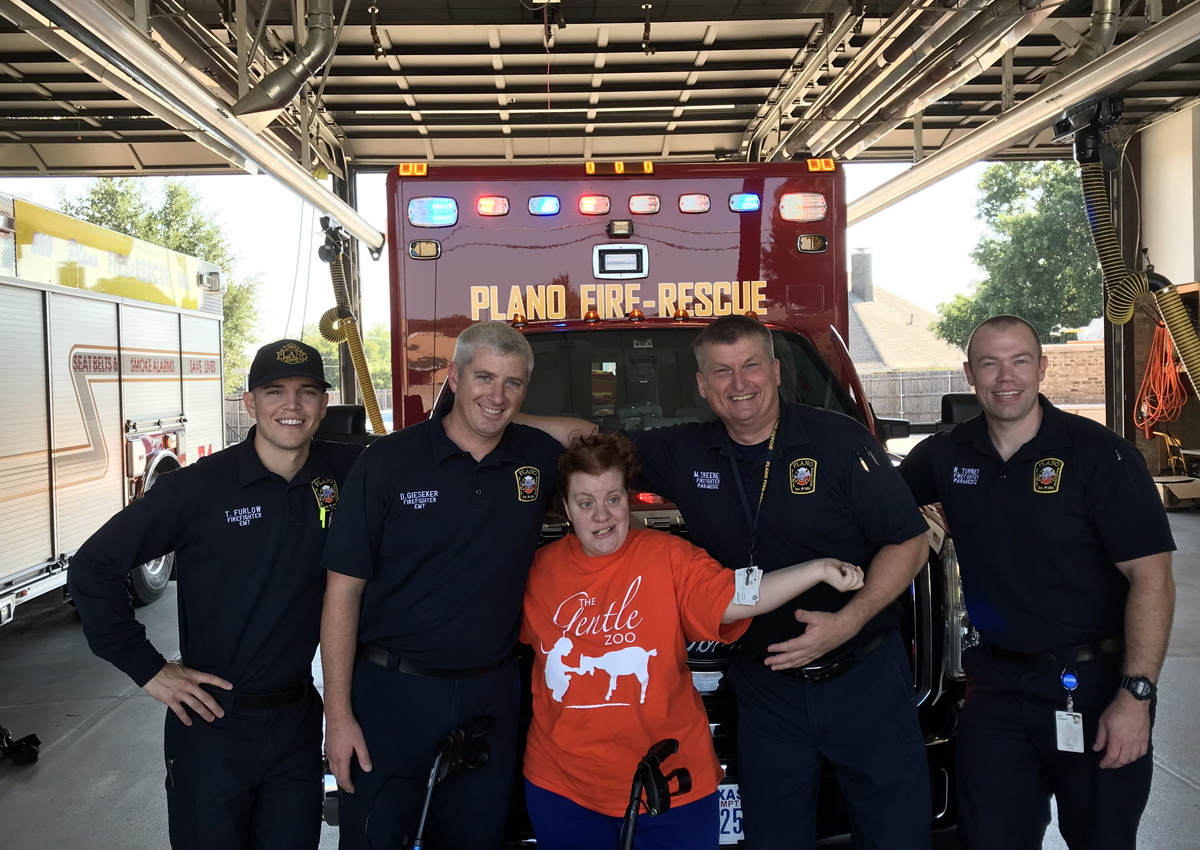 Four firemen in uniform smile while standing in front of a fire truck with a young adult.