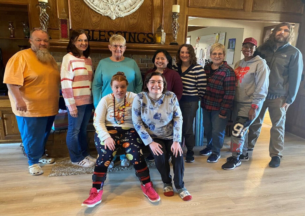 Eight adults stand in front of a fireplace. Two young adults sit in front of them.