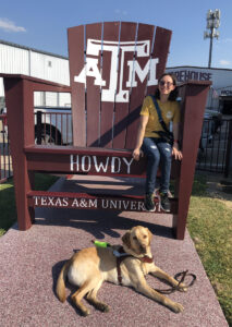 A young adult sits in an oversized adirondack style chair painted with the Texas A&M logo while her guide dog, Finn, rests on the ground below.