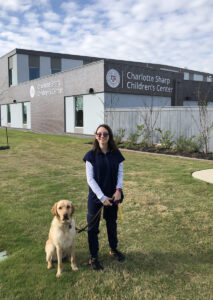 A young adult in navy work scrubs stands outside her work building with her guide dog.