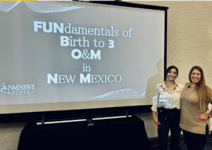 Two women stand next to a screen with the words “FUNdamentals of Birth to 3 O&M in New Mexico” written on it.