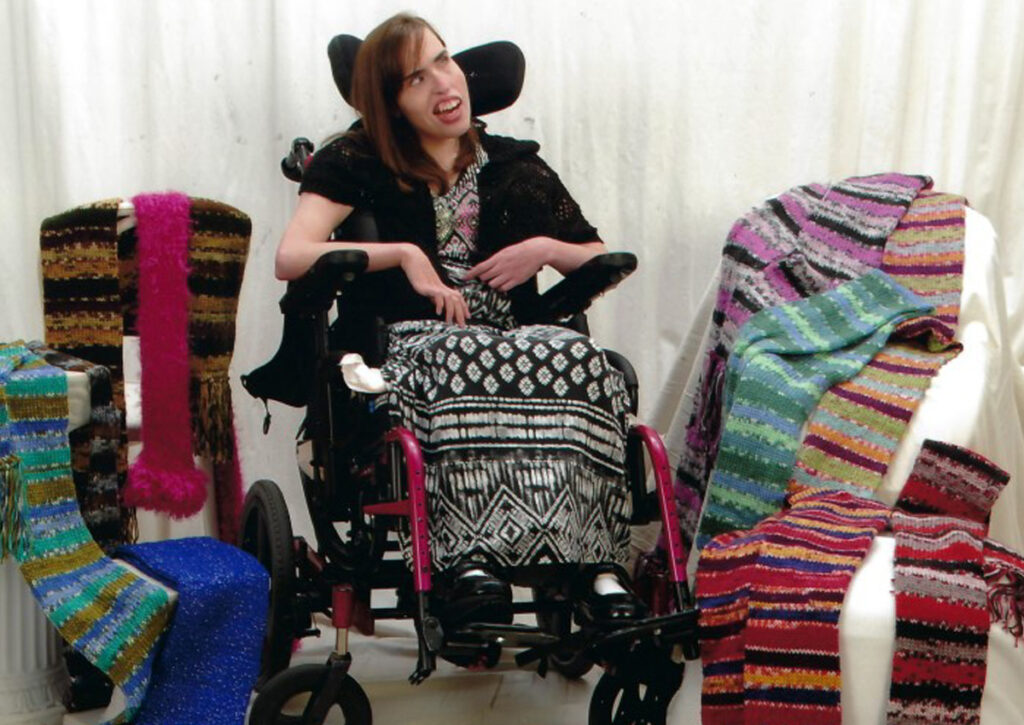 A young woman sitting in a pink wheelchair is surrounded by multiple colorful knitted scarves draped across white stone pillars placed on both sides of her chair.