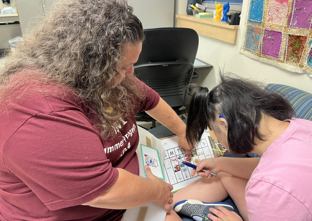 Un adolescente con las piernas dobladas en un sofá en un aula bien iluminada. El adulto que acompaña al lector sostiene abierto un libro personalizado en una página con un calendario semanal y señala una imagen en la columna del miércoles.
