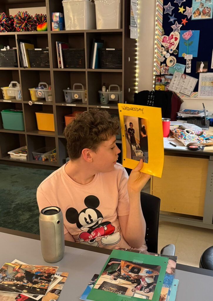 Un adolescente está sentado a una mesa y mira un trozo de papel amarillo brillante con la palabra “Miércoles” escrita encima de una foto de dos personas realizando una entrega. Sobre la mesa, frente a él, hay páginas personalizadas adicionales, y detrás del estudiante se ve un camino despejado hacia estantes bien organizados con contenedores de materiales.
