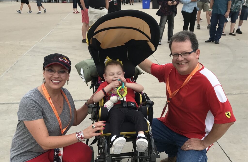 Mamá y papá, fotografiados con Abbey, sonríen mientras hacen una pausa en la propiedad de un evento de carreras de autos.