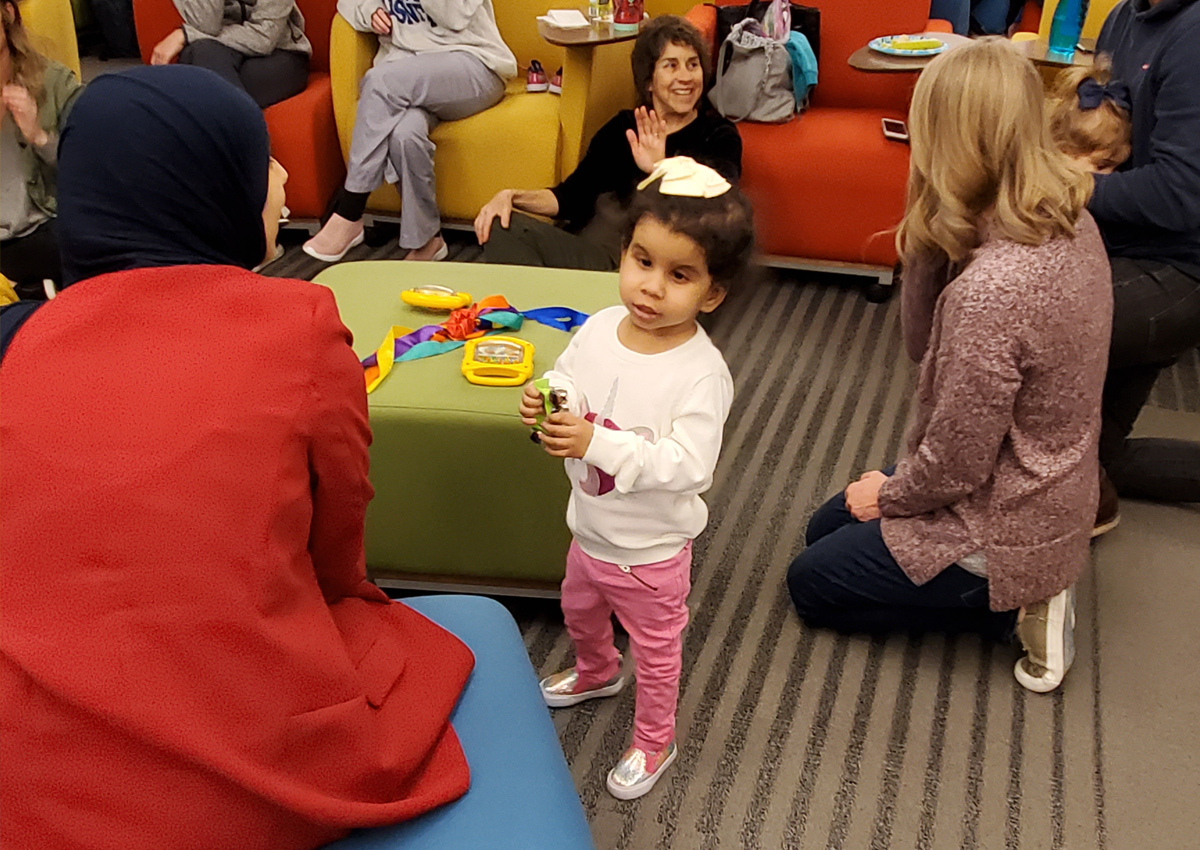 Un niño pequeño que sostiene una pulsera de cascabeles se encuentra en una concurrida sala de conferencias rodeado de adultos sentados en el suelo y en sillas acolchadas.