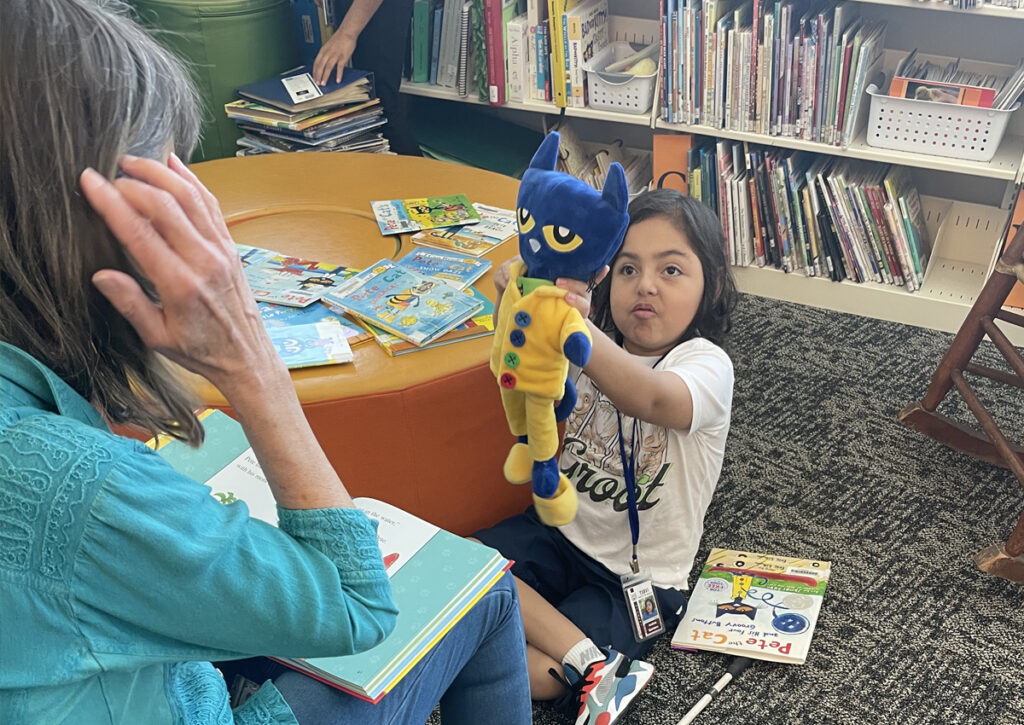 A child sitting on the floor of a library holds up a stuffed “Pete the Cat” doll to an adult who is reading to and seated across from her. “Pete the Cat” books are on the floor.