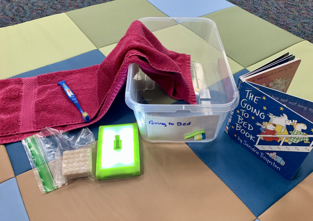 Book box for “The Going to Bed Book”, a commercial board book. An open plastic bin contains a red towel, a blue toothbrush, a plastic baggie containing a small soap bar, and a light switch mounted on a piece of foamboard.