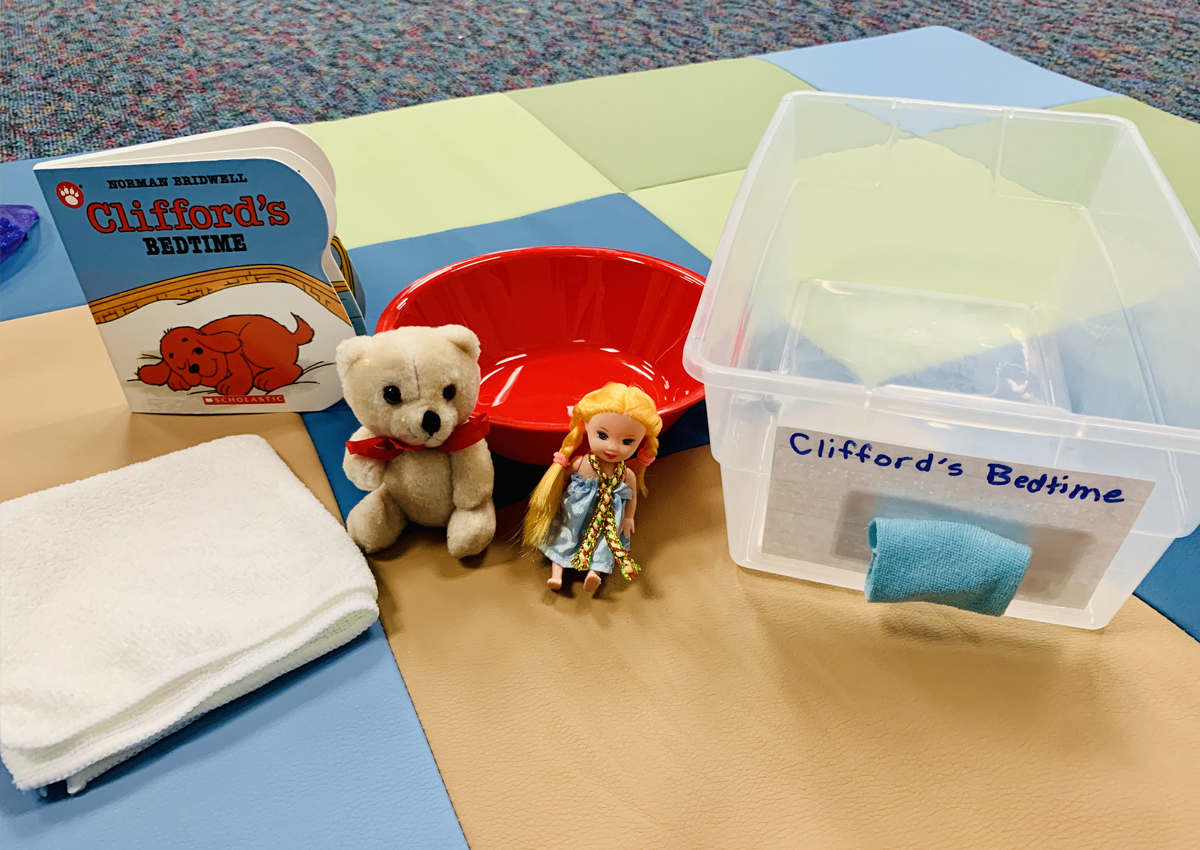 Book box for Clifford's Bedtime, a commercial board book. An open plastic bin has a label and a tactile symbol affixed to the side. Items for the story include a soft cloth, a small teddy bear, a small doll in a nightgown, and a red bowl.