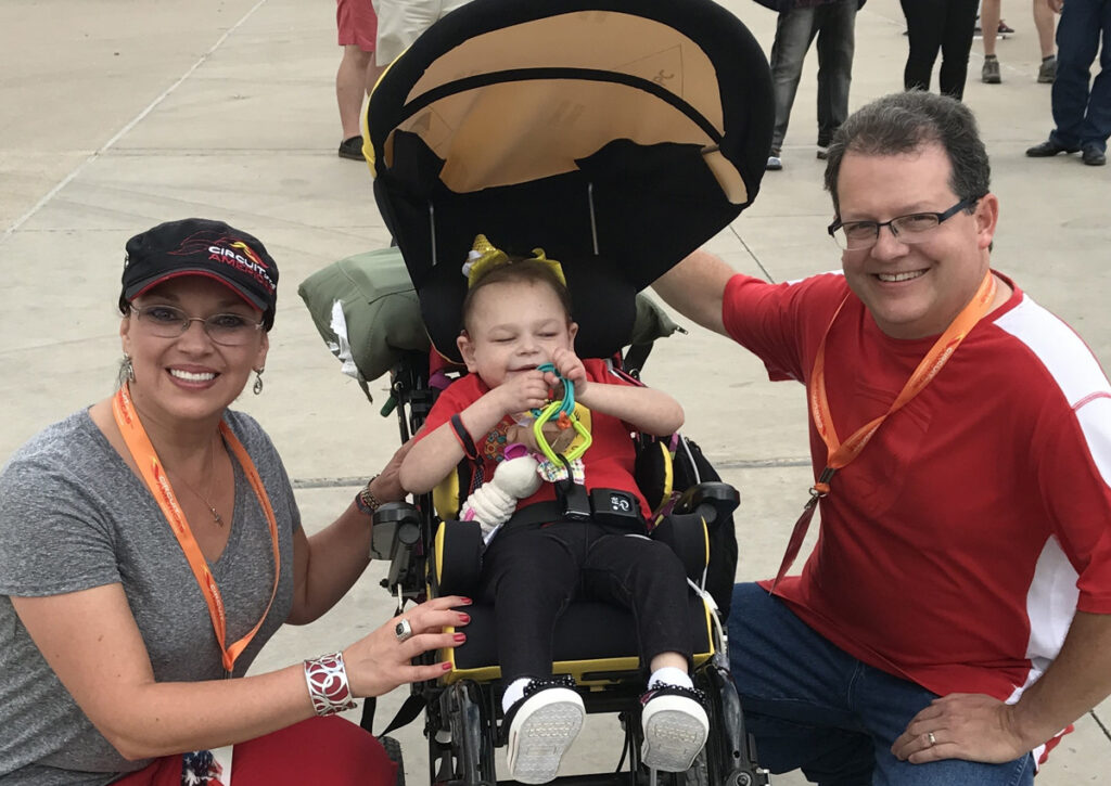 Mom and Dad, pictured with Abbey, are smiling as they pause on the grounds of a car racing event.