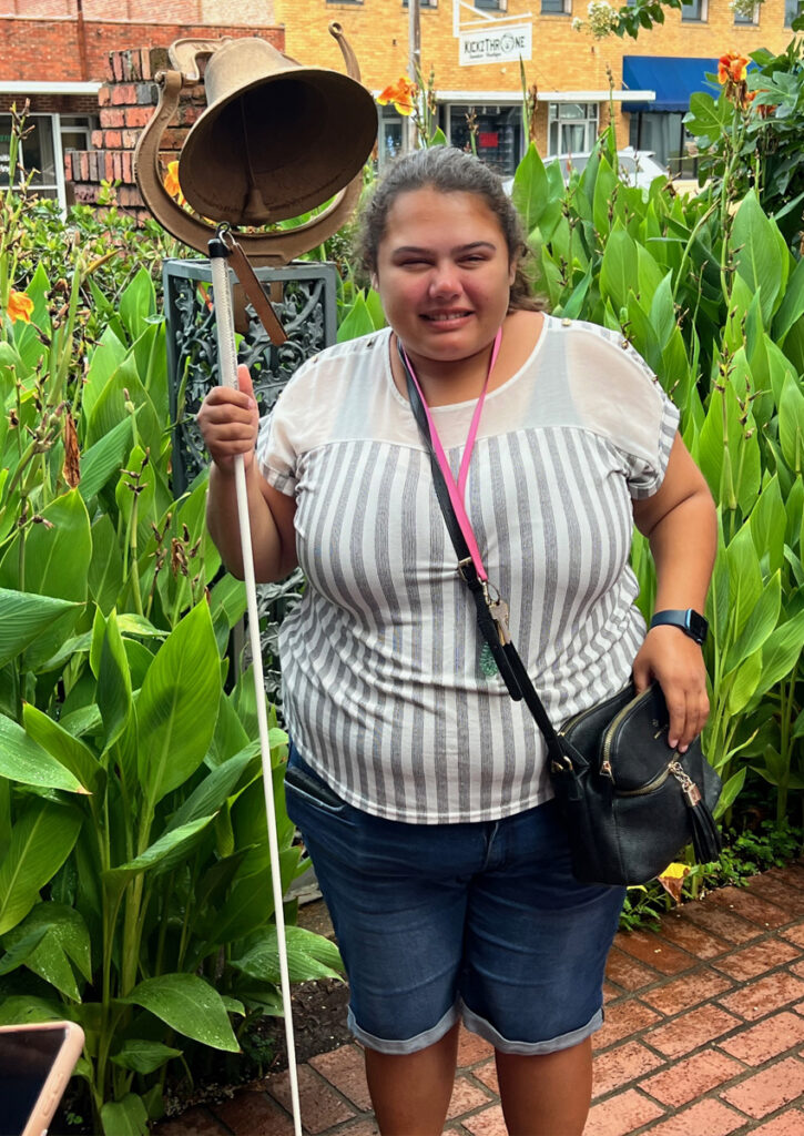 An adult with white cane stands outside in a garden next to a historic brass bell on a pedastal.