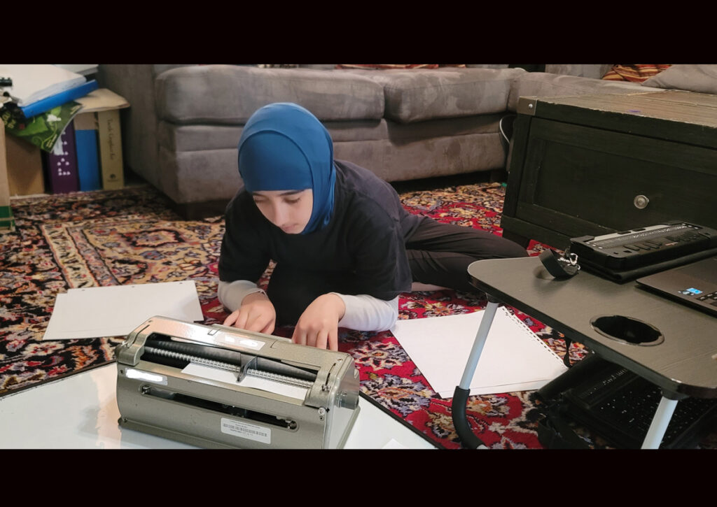 A student writes on a Perkins braillewriter while on the floor in her living room. A laptop and a refreshable braille display are to her left.