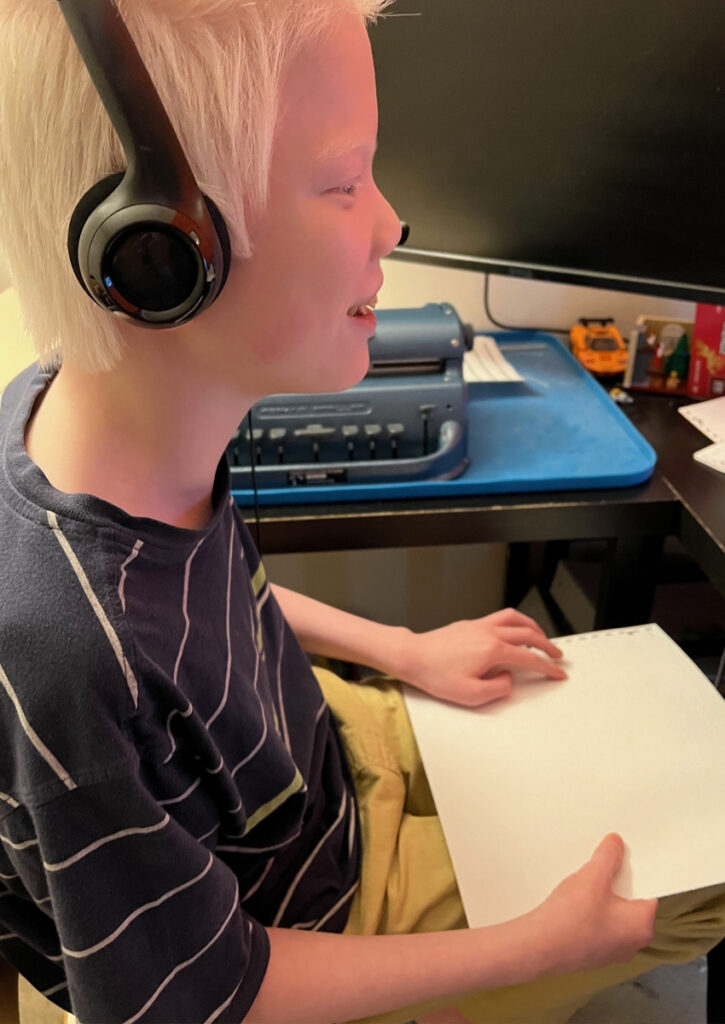 A student smiles as he reads a braille math problem from a page on his lap. The Perkins Braillewriter is next to him.