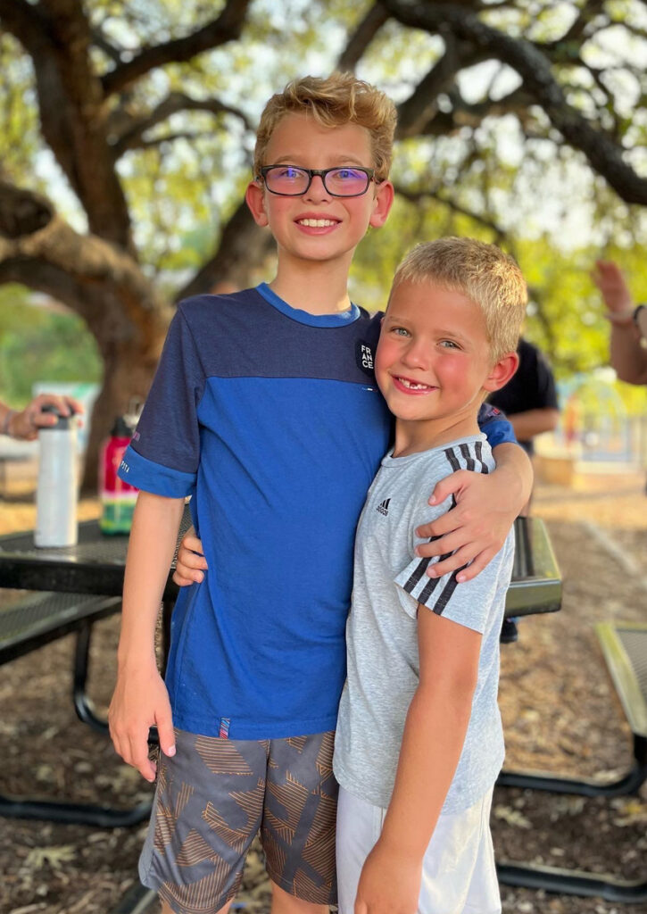 Two young boys smile and embrace while standing under a large shade tree.