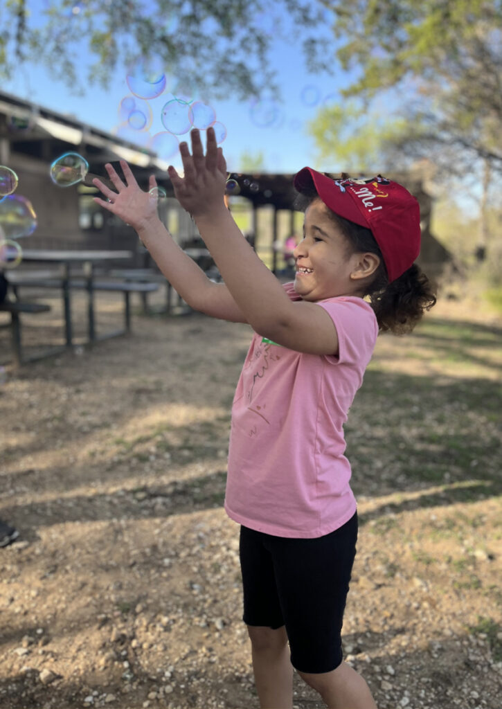 A young girl reaches up toward floating bubbles outdoors.