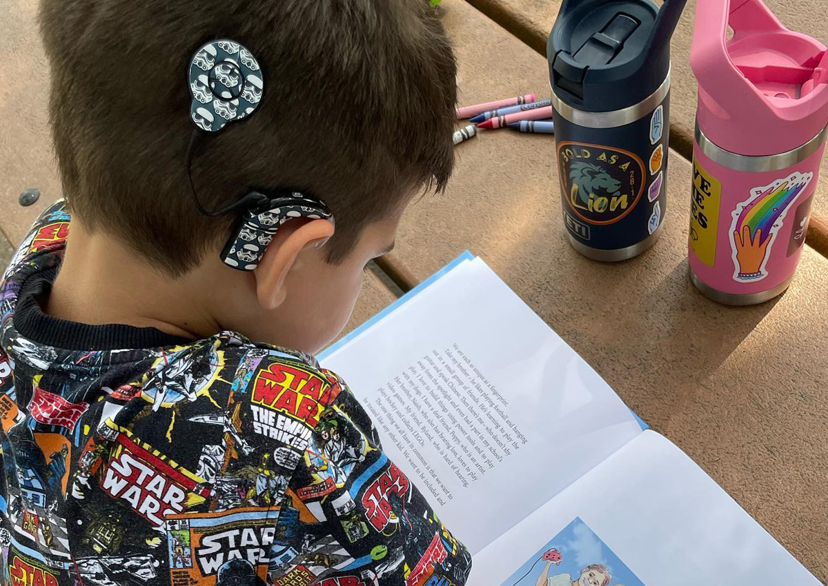 A boy with a Star Wars-themed cochlear implant sits at a table reading a book.