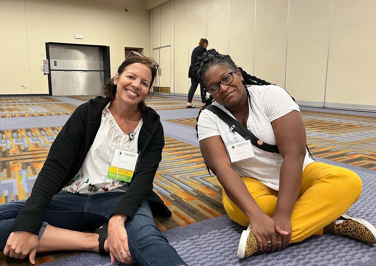 Two women sit cross-legged on a patterned carpet in a large conference hall.