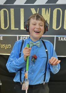 Wearing a bow tie, suspenders, and headphones, and holding his cane in front of him, Eddie laughs broadly in front of a sign about the school prom.