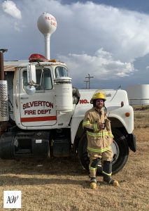 An adult in full firefighter gear stands next to a fire truck labeled Silverton Fire Department in an open field.