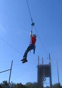 A young adult wearing a harness and safety helmet hangs from the zipline.
